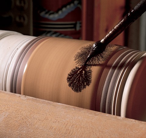 Detail show of the side of a hollow ceramic vessel on a lathe being decorated with black mocha tea.